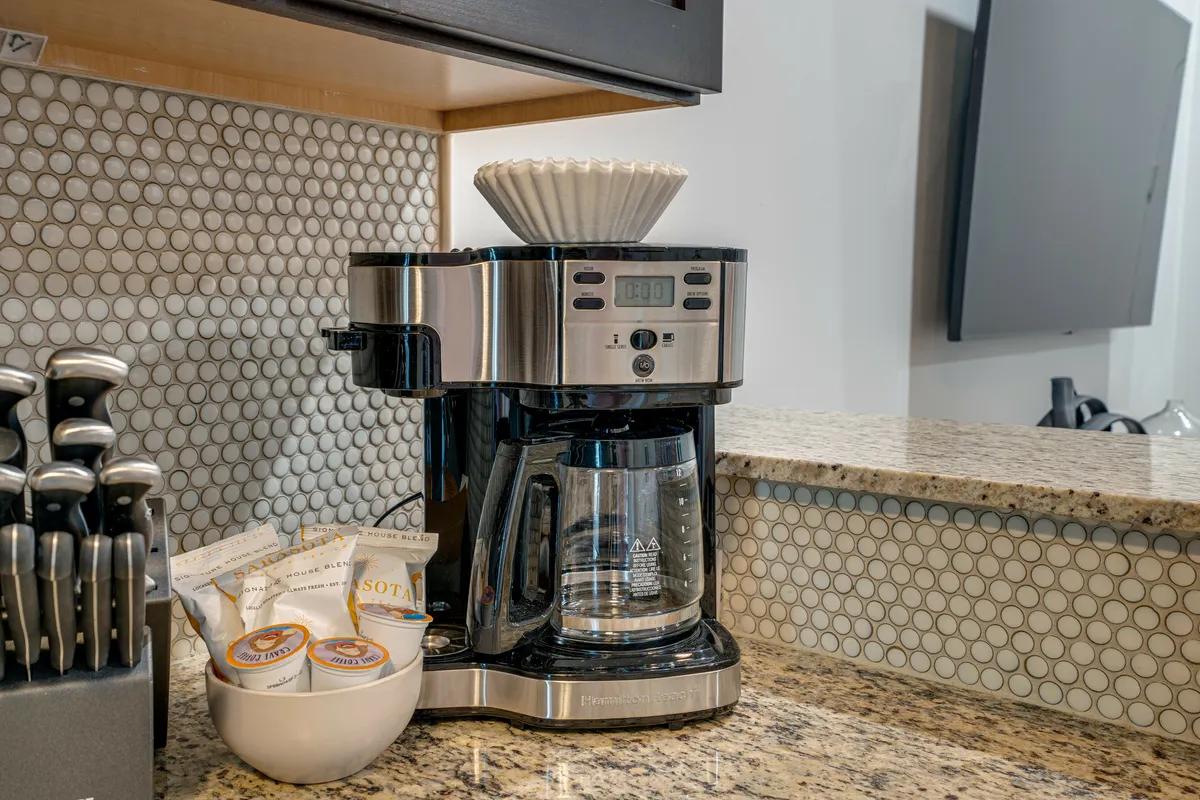 A close-up of a granite kitchen counter displays a coffee maker, coffee pods, a knife set, decorative backsplash, and other kitchen essentials. The setting is both stylish and functional, capturing the essence of hotel-style starter kits for guests.