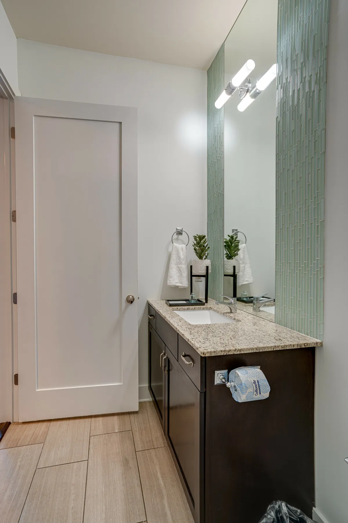 A modern bathroom with a granite countertop, large mirror, green glass tile backsplash, and contemporary fixtures. The design balances functionality with upscale, spa-like details.