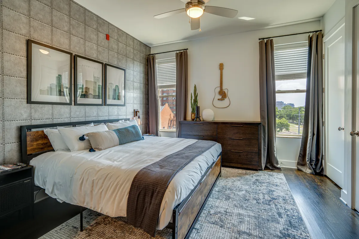A serene bedroom featuring a bed adorned with decorative pillows, artistic wall decor, a guitar mounted on the wall, and a dresser topped with green plants. The scene exudes a peaceful, inviting atmosphere suited for rest and relaxation.
