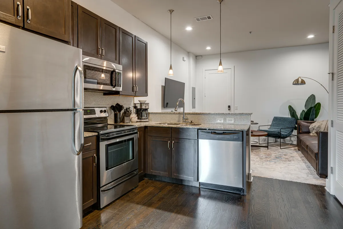 This image features a modern, cozy open-concept living and kitchen area. Stainless steel appliances and granite countertops stand out in the kitchen, while the living space offers comfortable seating perfect for relaxing or gathering with friends. Modern fixtures and a welcoming atmosphere make the space feel both practical and inviting.