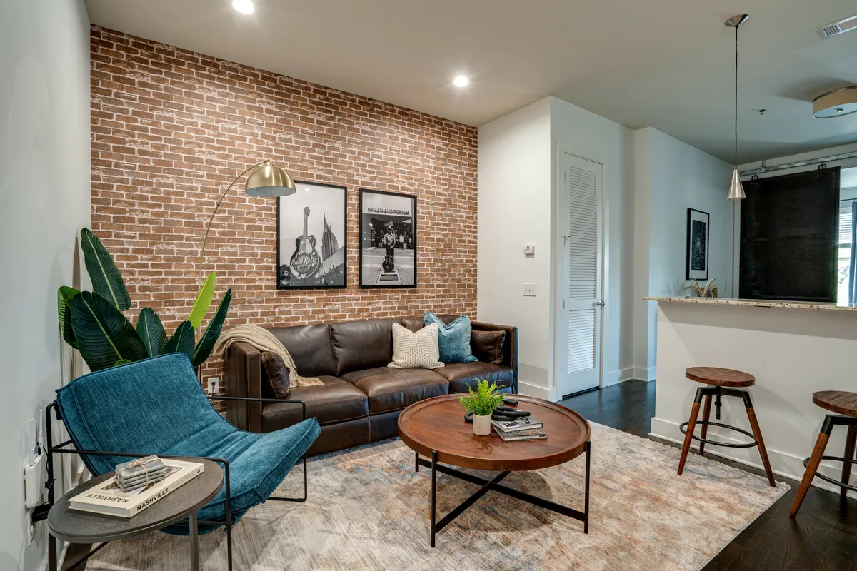 The hero image showcases a stylish open-concept living area featuring a striking exposed brick wall, a plush leather sofa, a modern blue accent chair, a sleek coffee table, lush green plants, and curated framed wall art. The scene is bathed in natural light, exuding a cozy yet contemporary atmosphere perfect for relaxation or socializing.